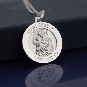 May include: Silver pendant with a depiction of Saint Joseph holding baby Jesus. The pendant is round and has the words "Saint Joseph Pray For Us" inscribed around the edge.