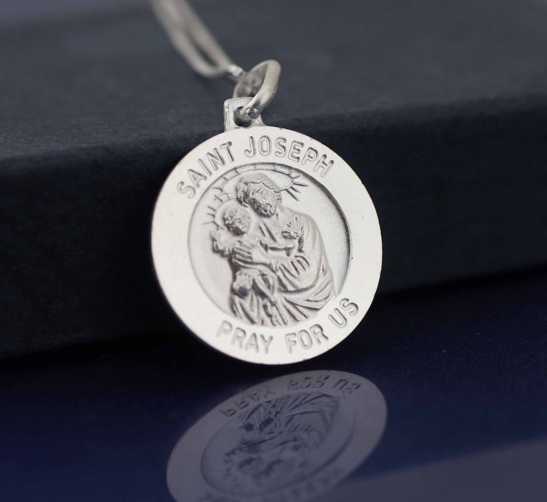 May include: Silver pendant with a depiction of Saint Joseph holding baby Jesus. The pendant is round and has the words "Saint Joseph Pray For Us" inscribed around the edge.
