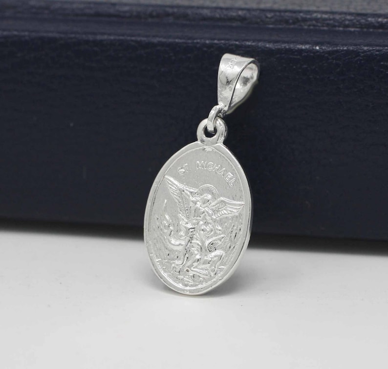 May include: Silver pendant featuring a detailed image of Saint Michael the Archangel with the words "St. Michael" inscribed around the oval-shaped pendant.