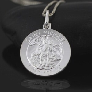 May include: Silver pendant necklace featuring a round charm with a depiction of Saint Michael the Archangel. The charm has the words "Saint Michael Pray For Us" inscribed around the edge.