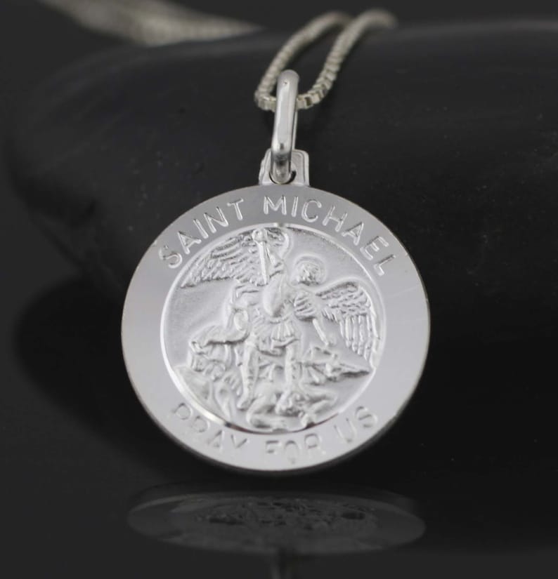 May include: Silver pendant necklace featuring a round charm with a depiction of Saint Michael the Archangel. The charm has the words "Saint Michael Pray For Us" inscribed around the edge.
