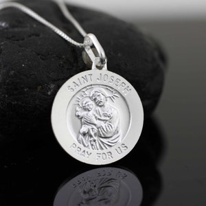 May include: Silver pendant necklace featuring a round medallion with a raised image of Saint Joseph holding baby Jesus. The medallion is inscribed with "Saint Joseph Pray For Us".