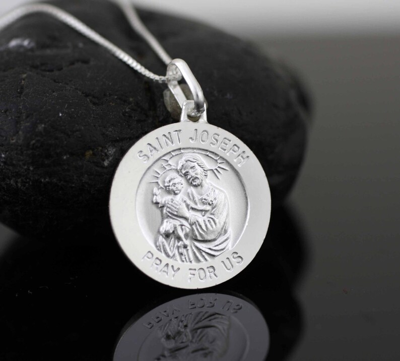 May include: Silver pendant necklace featuring a round medallion with a raised image of Saint Joseph holding baby Jesus. The medallion is inscribed with "Saint Joseph Pray For Us".