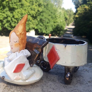May include: A ceramic figurine of a person with orange hair wearing a white and red costume, sitting on a white ceramic base with a red diamond. The figurine is part of a larger ceramic set, with a separate white ceramic lid with a red diamond and three legs.