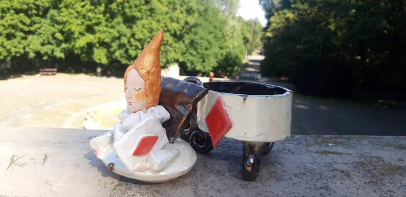 May include: A ceramic figurine of a person with orange hair wearing a white and red costume, sitting on a white ceramic base with a red diamond. The figurine is part of a larger ceramic set, with a separate white ceramic lid with a red diamond and three legs.