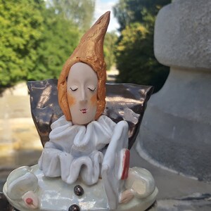 May include: A ceramic figurine of a person with a pointed hat, wearing a white ruffled collar and a brown and gold cape. The figurine is holding a red and white object in its hand.
