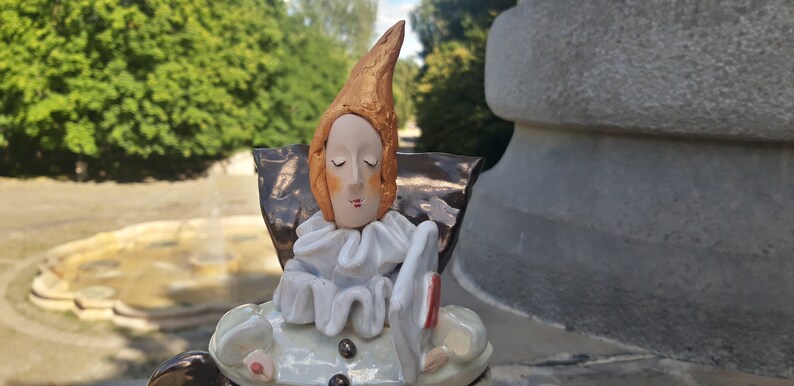 May include: A ceramic figurine of a person with a pointed hat, wearing a white ruffled collar and a brown and gold cape. The figurine is holding a red and white object in its hand.