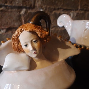 May include: A porcelain figurine of a woman with a crown, wearing a white dress and holding a pillow. The figurine is painted with gold accents and has a detailed facial expression.