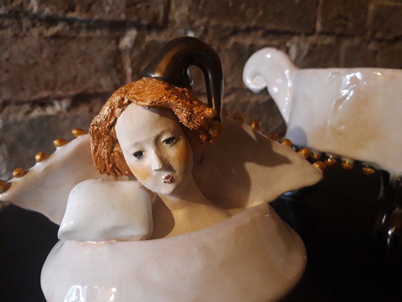 May include: A porcelain figurine of a woman with a crown, wearing a white dress and holding a pillow. The figurine is painted with gold accents and has a detailed facial expression.