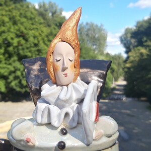May include: A ceramic figurine of a person with a pointed hat, wearing a white ruffle collar and holding a white plate. The figurine is painted with shades of brown, white, and gold.