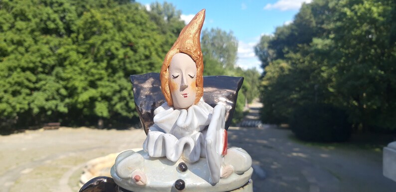 May include: A ceramic figurine of a person with a pointed hat, wearing a white ruffle collar and holding a white plate. The figurine is painted with shades of brown, white, and gold.