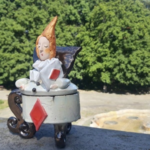 May include: A ceramic figurine of a person sitting in a chair shaped like a playing card. The chair is a diamond and the person is wearing a white and red costume. The figurine is sitting on a stone surface with a fountain in the background.