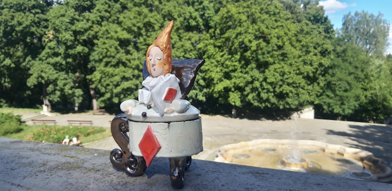 May include: A ceramic figurine of a person sitting in a chair shaped like a playing card. The chair is a diamond and the person is wearing a white and red costume. The figurine is sitting on a stone surface with a fountain in the background.