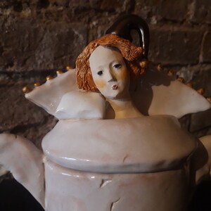 May include: A white ceramic teapot with a whimsical design featuring a woman's head with red hair and a black hat. The teapot has a white lid and a gold trim.