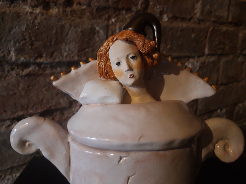 May include: A white ceramic teapot with a whimsical design featuring a woman's head with red hair and a black hat. The teapot has a white lid and a gold trim.