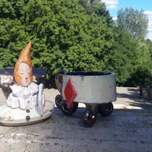 May include: A ceramic figurine of a jester with a white ruff and a red diamond on a white ceramic cup with wheels.