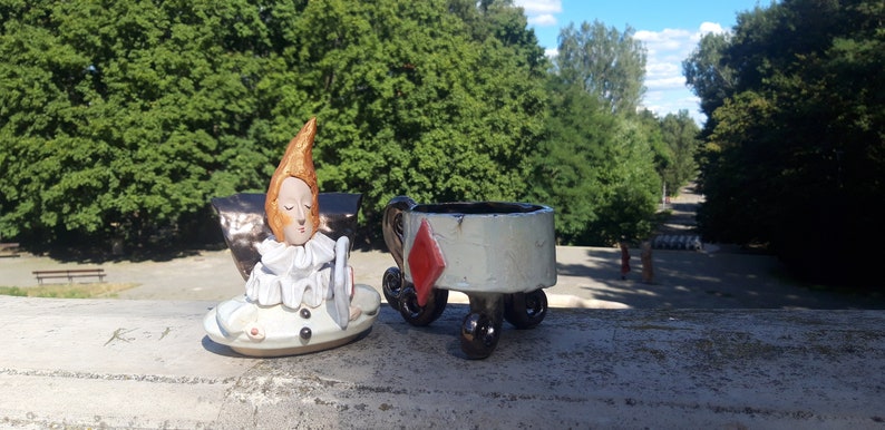 May include: A ceramic figurine of a jester with a white ruff and a red diamond on a white ceramic cup with wheels.