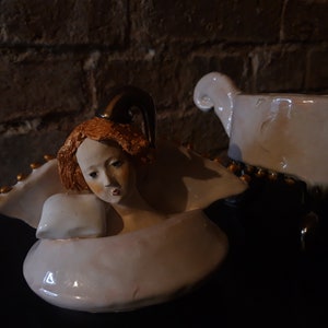 May include: A white ceramic figurine of an angel with a gold halo and wings. The angel has red hair and is lying down with its head resting on a pillow. The figurine is on a black surface.