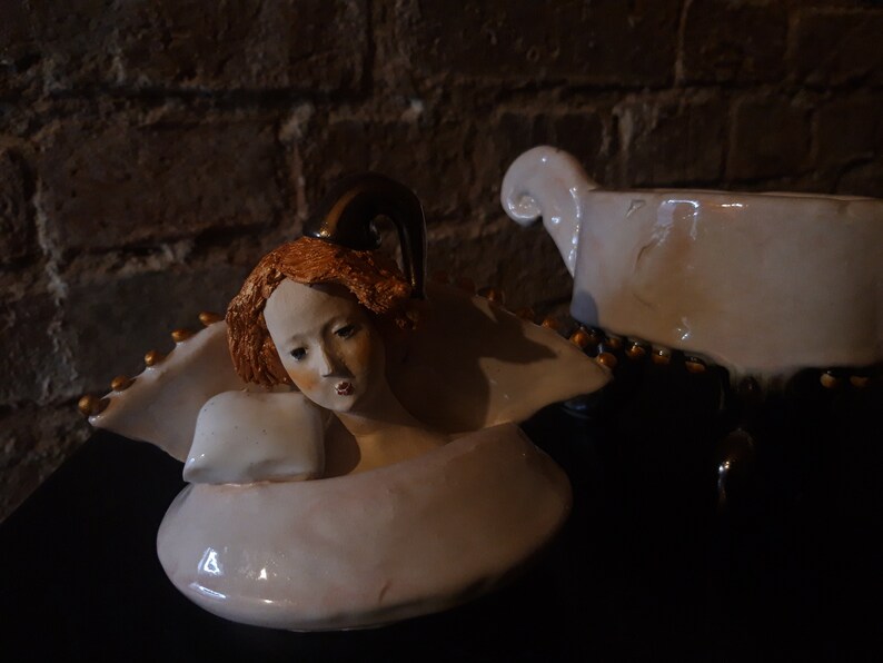 May include: A white ceramic figurine of an angel with a gold halo and wings. The angel has red hair and is lying down with its head resting on a pillow. The figurine is on a black surface.