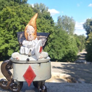 May include: A ceramic figurine of a person with a pointed hat, wearing a white and black costume, sitting on a chair with a red diamond design. The figurine is sitting on a stone surface with a green background.