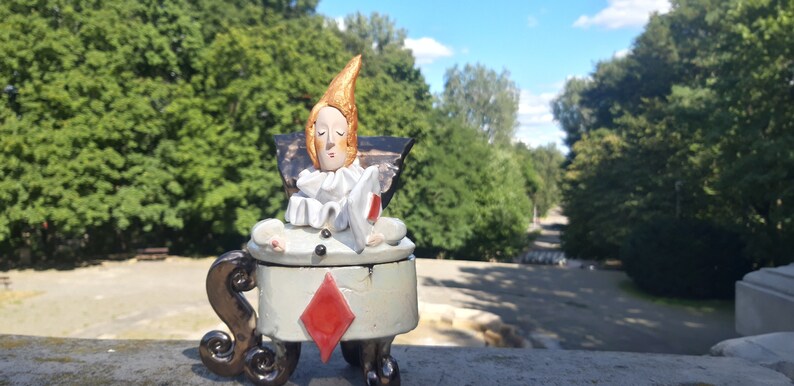 May include: A ceramic figurine of a person with a pointed hat, wearing a white and black costume, sitting on a chair with a red diamond design. The figurine is sitting on a stone surface with a green background.