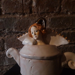 May include: A white ceramic teapot with a sculpted woman's head as the lid. The woman has red hair and is wearing a white dress with gold accents.