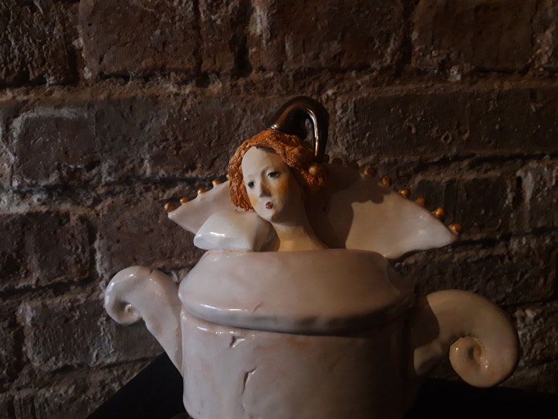 May include: A white ceramic teapot with a sculpted woman's head as the lid. The woman has red hair and is wearing a white dress with gold accents.