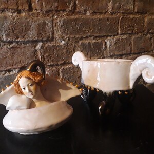 May include: A white ceramic lidded dish with a sculpted angel figure on the lid. The dish has three black legs with gold accents and a white glaze. The angel has a gold crown and a white dress.