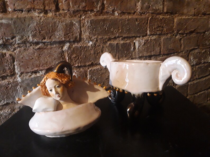May include: A white ceramic lidded dish with a sculpted angel figure on the lid. The dish has three black legs with gold accents and a white glaze. The angel has a gold crown and a white dress.