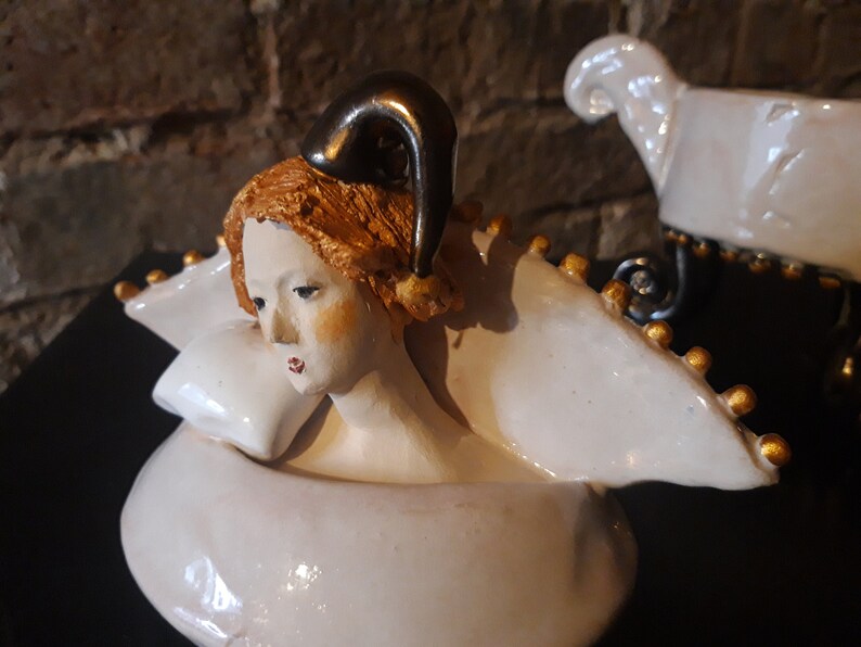 May include: A white ceramic sculpture of a woman with a gold crown and a gold horn on her head. The sculpture is resting on a white pillow.