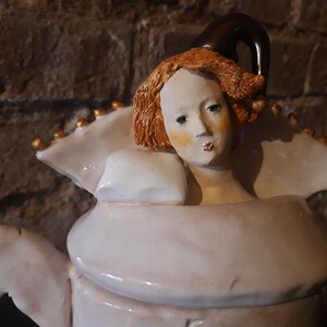May include: A white ceramic teapot with a sculpted woman's head as the lid. The woman has red hair and a white dress. The teapot has gold accents.