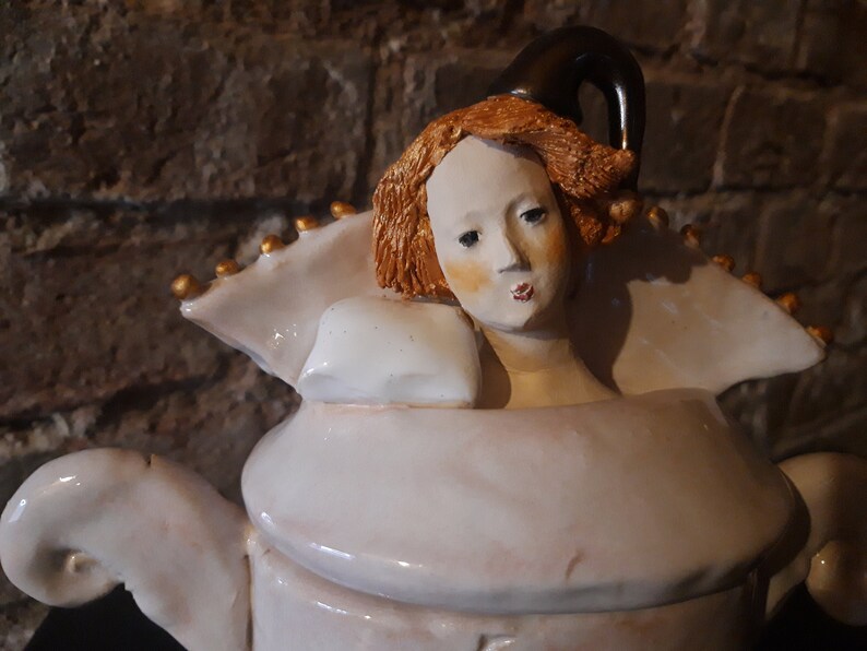 May include: A white ceramic teapot with a sculpted woman's head as the lid. The woman has red hair and a white dress. The teapot has gold accents.