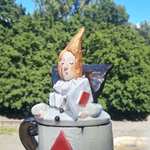 May include: A ceramic teapot with a whimsical design. The lid features a sculpted figure of a person with a pointed hat, wearing a white ruff and holding a red playing card. The teapot is painted in shades of blue, white, and red.