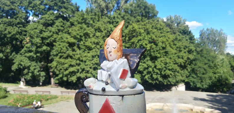 May include: A ceramic teapot with a whimsical design. The lid features a sculpted figure of a person with a pointed hat, wearing a white ruff and holding a red playing card. The teapot is painted in shades of blue, white, and red.