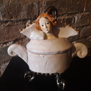 May include: A white ceramic teapot with a whimsical design featuring a woman's head with red hair and a black hat. The teapot has gold accents and sits on three silver legs.