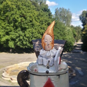 May include: A ceramic figurine of a person with a gold crown and white collar, sitting on a white ceramic box with a red diamond design. The box has black, curly legs.