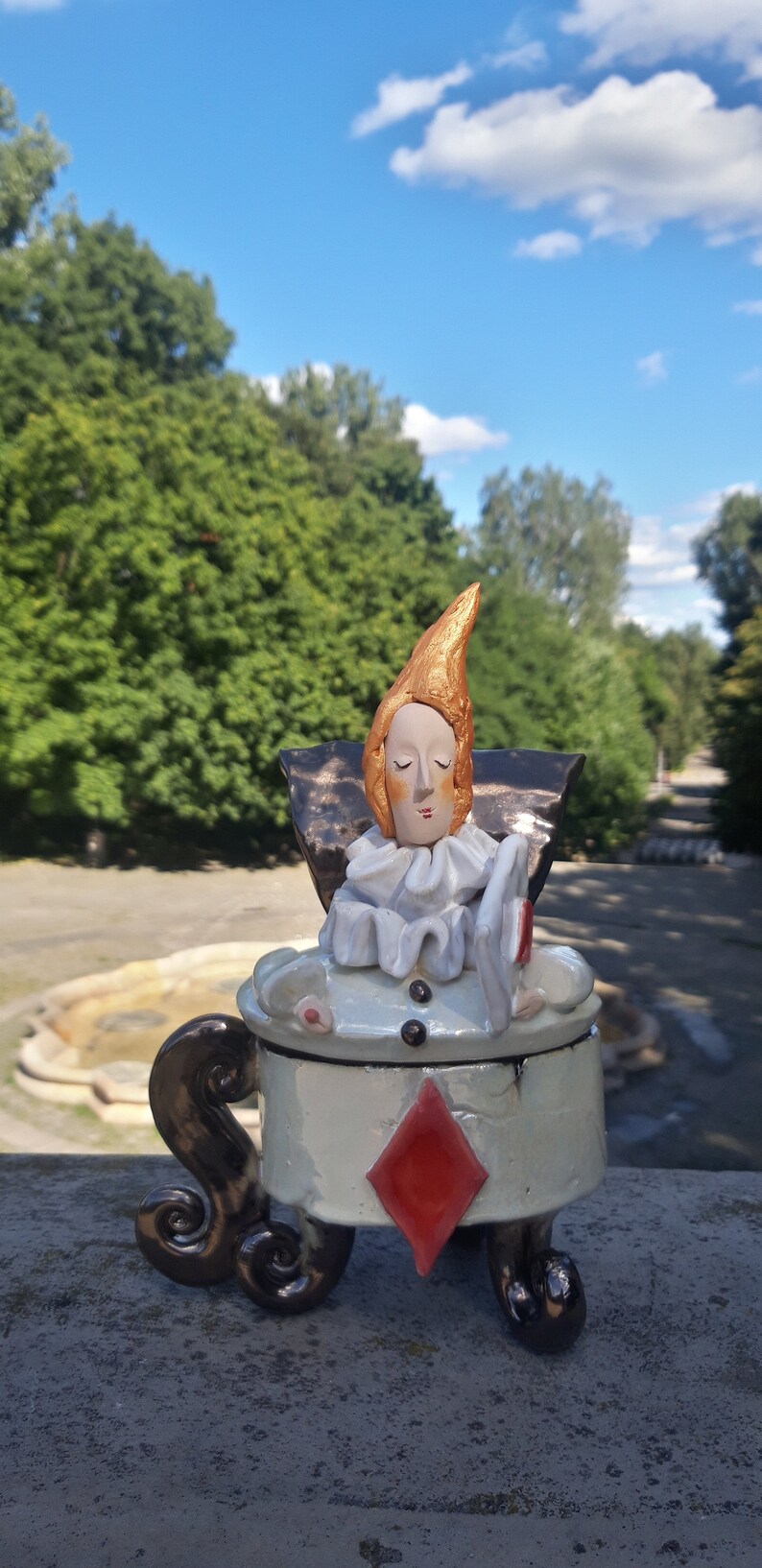 May include: A ceramic figurine of a person with a gold crown and white collar, sitting on a white ceramic box with a red diamond design. The box has black, curly legs.