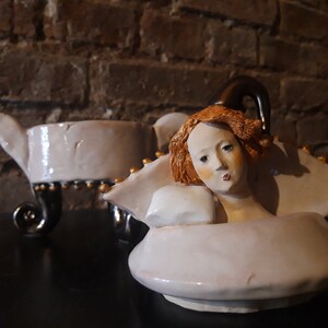 May include: A white ceramic teapot with a gold trim and a sculpted woman's head with red hair. The teapot has a spout shaped like a swan's neck and a lid with a finial.