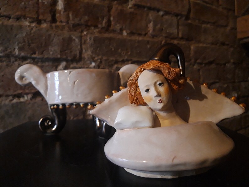 May include: A white ceramic teapot with a gold trim and a sculpted woman's head with red hair. The teapot has a spout shaped like a swan's neck and a lid with a finial.