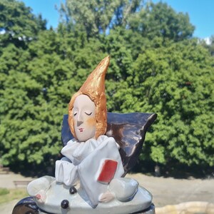 May include: A ceramic figurine of a person with a pointed hat, wearing a white and red costume. The figurine is sitting on a black base and has a red heart in its hand.