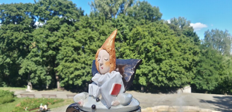 May include: A ceramic figurine of a person with a pointed hat, wearing a white and red costume. The figurine is sitting on a black base and has a red heart in its hand.