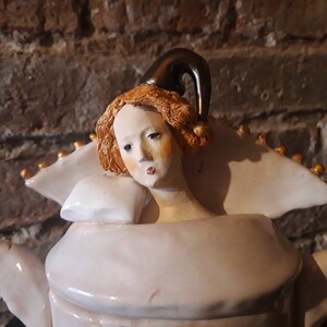May include: A white ceramic teapot with a gold trim and a woman's head with a jester's hat on top. The woman has red hair and is wearing a white collar.