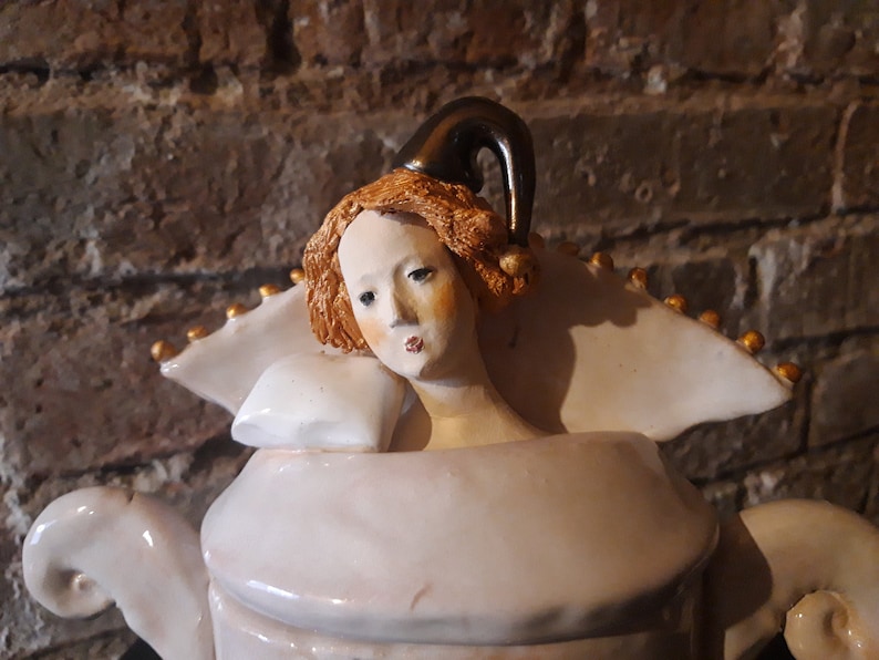 May include: A white ceramic teapot with a gold trim and a woman's head with a jester's hat on top. The woman has red hair and is wearing a white collar.