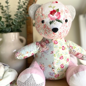 May include: A stuffed bear made from patchwork fabric featuring various floral patterns in shades of pink, green, and blue. The bear has grey ears, black button eyes, a black nose, and three small pink buttons on its chest. The left foot displays the text BORN IN 2019.