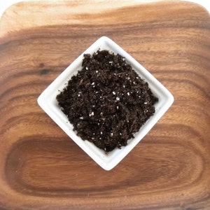 Puede incluir: Un plato cuadrado de cer&aacute;mica blanca lleno de tierra marr&oacute;n oscura, salpicada de gr&aacute;nulos blancos. El plato est&aacute; sobre una bandeja cuadrada de madera con un patr&oacute;n de vetas de madera natural. El fondo es blanco.
