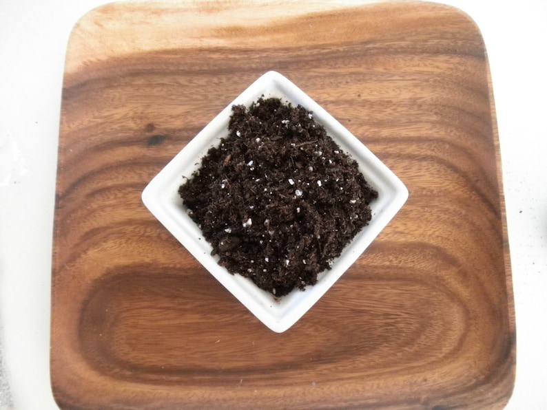 Puede incluir: Un plato cuadrado de cer&aacute;mica blanca lleno de tierra marr&oacute;n oscura, salpicada de gr&aacute;nulos blancos. El plato est&aacute; sobre una bandeja cuadrada de madera con un patr&oacute;n de vetas de madera natural. El fondo es blanco.