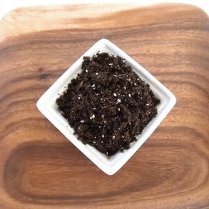 Puede incluir: Un plato cuadrado de cer&aacute;mica blanca lleno de tierra marr&oacute;n oscuro, salpicado de part&iacute;culas blancas, se encuentra sobre una tabla de madera. La tabla tiene un patr&oacute;n de vetas de madera natural y un color marr&oacute;n c&aacute;lido. La tierra parece lista para plantar.