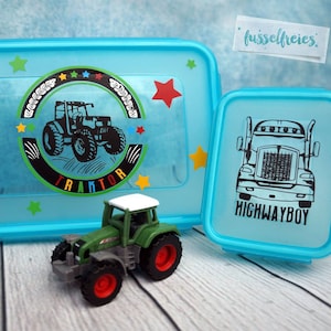 Könnte beinhalten: Zwei blaue Lunchboxen mit einem Traktor- und einem LKW-Design. Die Lunchbox mit dem Traktor-Design hat ein transparentes Fenster und Sterne am Rand. Die Lunchbox mit dem LKW-Design hat die Worte "HIGHWAYBOY" auf der Vorderseite aufgedruckt. Ein kleiner grüner Spielzeugtraktor befindet sich im Vordergrund.