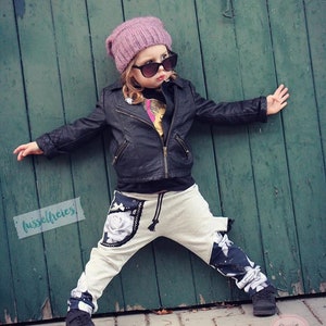 Puede incluir: Una persona joven que lleva una chaqueta de cuero negra, un gorro rosa y gafas de sol. Lleva un pantal&oacute;n blanco con estampado floral blanco y negro y zapatos negros. La persona est&aacute; de pie frente a una puerta de madera verde.