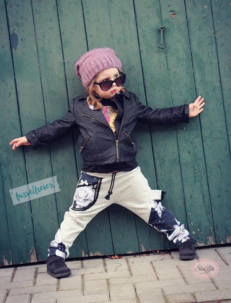 Puede incluir: Una persona joven que lleva una chaqueta de cuero negra, un gorro rosa y gafas de sol. Lleva un pantal&oacute;n blanco con estampado floral blanco y negro y zapatos negros. La persona est&aacute; de pie frente a una puerta de madera verde.